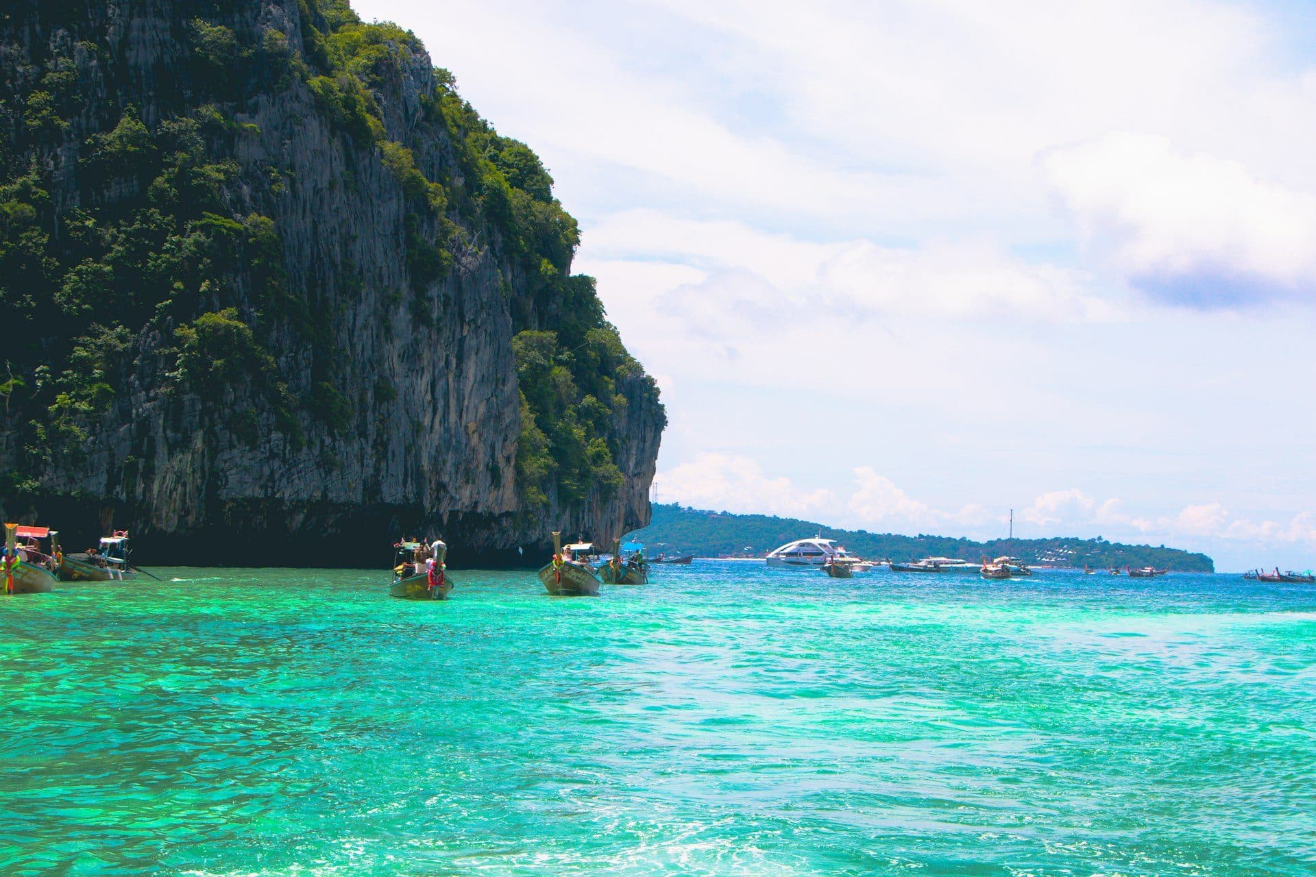 Longtail boats in turquoise water beside a lush green cliff in Thailand